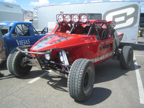 1990 Ford Bronco - red tube-chassis off-road race car numbered 102 with a four-light roof bar parked on pavement beside team haulers.