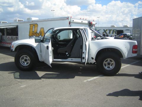 1990 Ford Bronco - white off-road prerunner pickup sits with both doors open in a parking lot beside enclosed team trailers.
