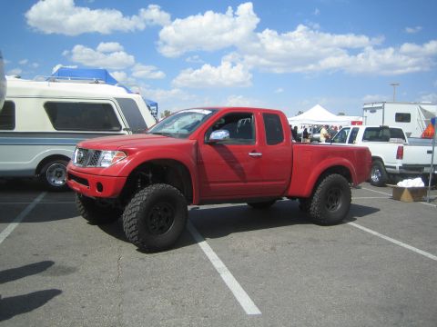 1990 Ford Bronco - red Nissan Frontier extended-cab pickup lifted on large off-road tires parked in a lot beside an RV and tents.