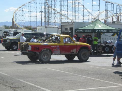 1990 Ford Bronco - yellow-and-red off-road race truck numbered 744 sits in a paved pit area with a roller coaster on the horizon.