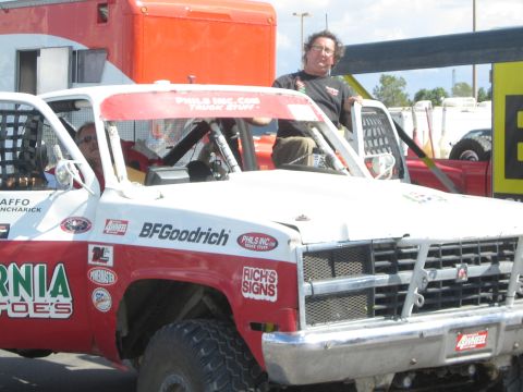 1990 Ford Bronco - man sits in a white-and-red BFGoodrich-sponsored off-road race truck covered in sponsor decals in a paved lot.
