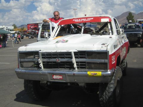 1990 Ford Bronco - Front view of a white-and-red off-road race truck with a tubular bumper as a man leans on its roof in a lot.