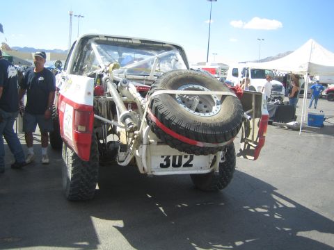 1990 Ford Bronco - Rear view of a white and red desert race truck carrying a spare tire on a tailgate carrier, marked number 302, parked in a race paddock.