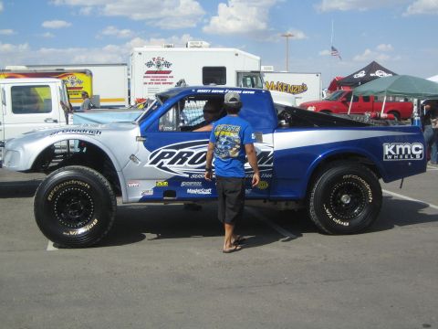 1990 Ford Bronco - blue and white PROAM-branded desert race truck on oversized BFGoodrich tires, with a person in a graphic shirt standing alongside in the pi…