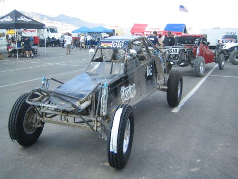 1990 Ford Bronco - black tube-frame single-seat off-road race buggy marked number 1616 sits in a paddock with tents, other buggies and a flag in the backgroun…