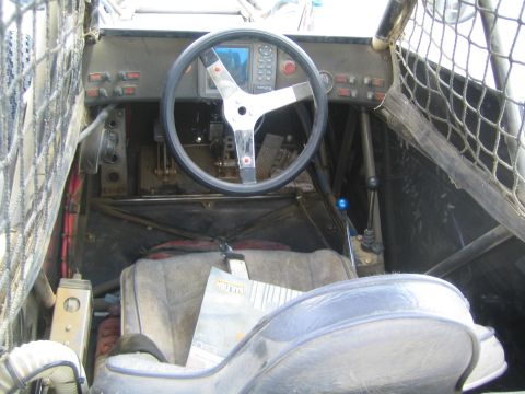 1990 Ford Bronco - Cockpit of an off-road race buggy with a three-spoke steering wheel, digital dash gauge, switch panels, netting and a worn bucket seat.