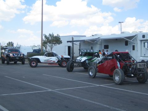 1990 Ford Bronco - lineup of off-road race buggies and trucks, including one numbered 1618, parked beside RVs and trailers in a paved staging lot.