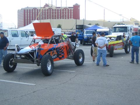 1990 Ford Bronco - orange and blue single-seat off-road race buggy with a raised rear wing sits in a parking lot as people gather among other race vehicles.