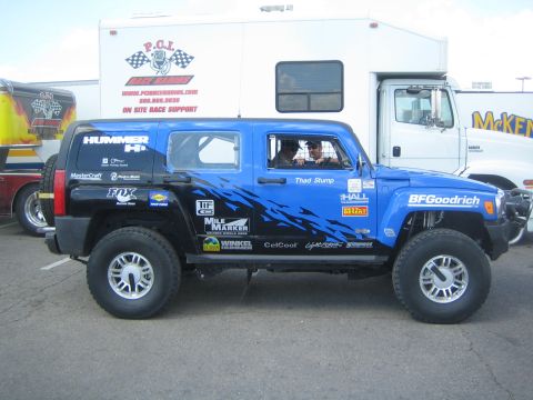 1990 Ford Bronco - Side view of a blue and black Hummer H3 desert race truck with HALL, BFGoodrich and other sponsor graphics, parked by a PCI support trailer.