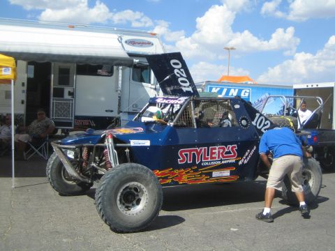 1990 Ford Bronco - dark blue off-road race truck number 1020 with Stylers Collision Repair graphics, hood up, as a crew member works on it in the pits.