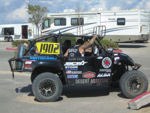 1990 Ford Bronco - black side-by-side UTV number 1902 with Desert Assassins sponsor graphics carrying two seated occupants, parked in front of trailers and an…
