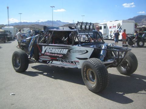 1990 Ford Bronco - black tube-frame off-road race buggy with Yochheim Racing graphics and a roof-mounted light bar parked in a paddock with mountains behind.
