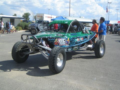1990 Ford Bronco - green ocean-themed off-road race buggy number 1006 on large knobby tires sits in a paddock as two people stand beside it.