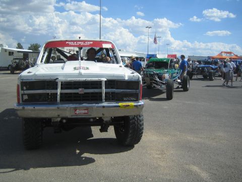 1990 Ford Bronco - Head-on view of a white lifted race truck with a chrome brush guard moving through a dusty, crowded off-road staging area.
