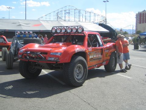 1990 Ford Bronco - orange off-road race truck with a roof-mounted light bar and Wide Open graphics parked near a roller coaster, with a man standing alongside.
