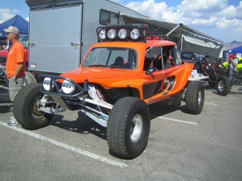 1990 Ford Bronco - orange and black tube-frame off-road race buggy with a row of PIAA driving lights parked in a paddock beside trailers.