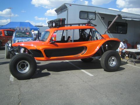 1990 Ford Bronco - glossy orange smooth-bodied off-road buggy with a roof storage rack and overhead lights parked beside a white RV in a paddock.