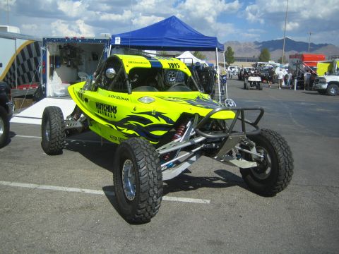 1990 Ford Bronco - bright yellow tube-frame off-road race buggy number 1913 with Hutchins and flame graphics parked under tents in a desert paddock.