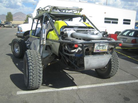 1990 Ford Bronco - Rear view of a yellow and silver off-road race buggy with prominent exhaust pipes and a license plate, parked among trucks and RVs.
