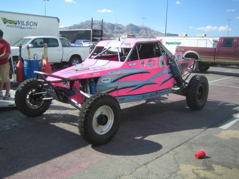 1990 Ford Bronco - pink and teal tribal-graphic off-road race buggy on large tires parked in a lot beside a Wilson Motorsports trailer with mountains behind.