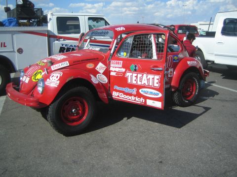 1990 Ford Bronco - red Volkswagen Beetle-bodied Class 5 desert race car covered in Tecate, SCORE and BFGoodrich sponsor decals sits in a paved pit lot.