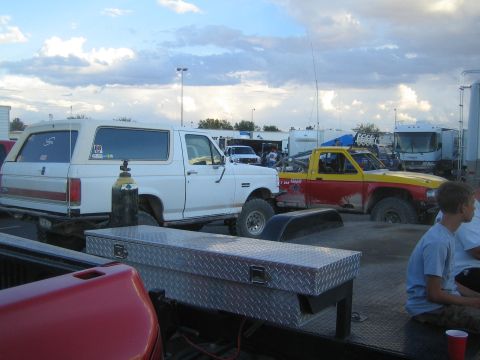 1990 Ford Bronco - pit scene showing a white Ford Bronco, a yellow-and-red off-road truck, diamond-plate toolboxes and a seated boy at dusk.