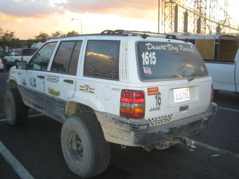 1990 Ford Bronco - Rear of a mud-spattered white Jeep Grand Cherokee marked Desert Dog Racing and number 1615, parked in a lot at sunset.