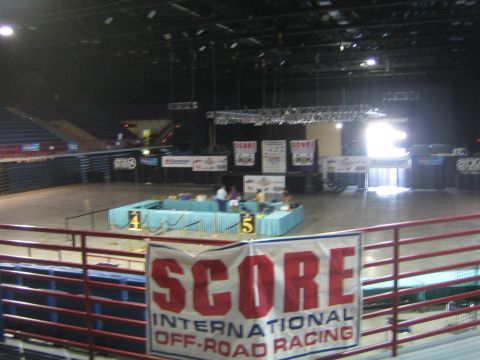 1990 Ford Bronco - Interior of a dark arena with staging tables and a large red SCORE International Off-Road Racing banner hanging on the front railing.