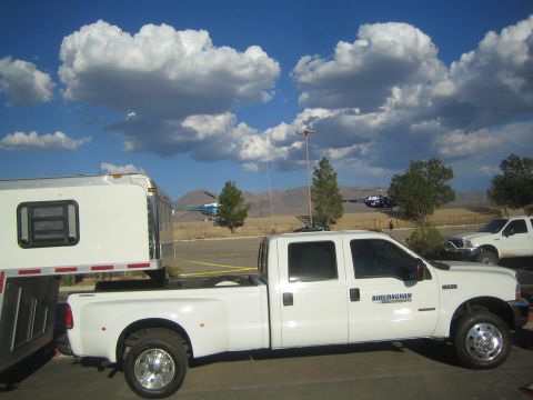 1990 Ford Bronco - white crew-cab dual-rear-wheel pickup hitched to a trailer, with two helicopters parked on dry desert ground under a cloudy sky.