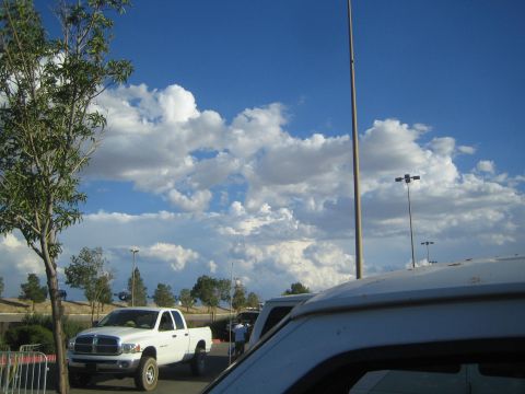 1990 Ford Bronco - white Dodge Ram pickup parked in a lot with tall light poles and a dramatic blue sky full of cumulus clouds.