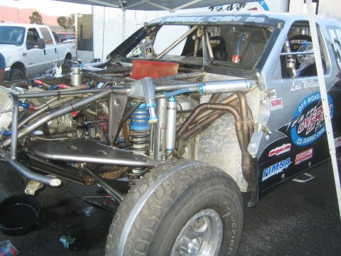 1990 Ford Bronco - Close-up of the exposed chromoly tube chassis, coilover shock and exhaust of a SCORE desert race car parked in a pit.