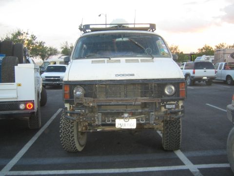 1990 Ford Bronco - Head-on view of a lifted white Dodge van riding on large off-road tires with a roof light bar, parked in a lot at dusk.