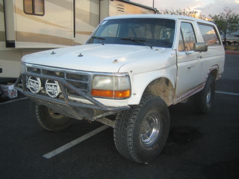 1990 Ford Bronco - white lifted Ford Bronco fitted with a tube brush guard and Hella driving lights parked beside a motorhome at dusk.
