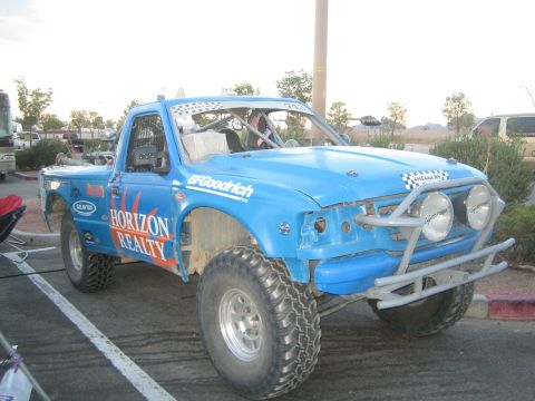 1990 Ford Bronco - blue single-cab off-road race truck lettered Horizon Realty with a tube front bumper and twin driving lights parked in a lot at dusk.