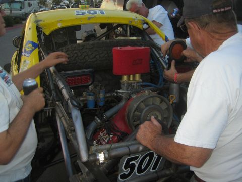 1990 Ford Bronco - Crew members work on the exposed rear-mounted engine of a yellow number 501 off-road race truck at dusk.