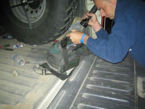 1990 Ford Bronco - man in a blue jacket uses a flashlight to inspect components while leaning from a truck bed at night.