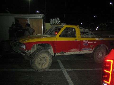 1990 Ford Bronco - yellow and red number 744 off-road race truck with roof light bar parked in a lot at night.