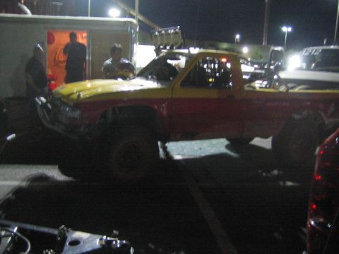1990 Ford Bronco - yellow and red off-road race truck with roof lights staged near an open enclosed trailer at night.