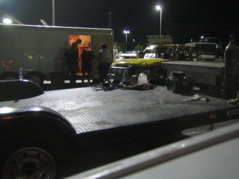 1990 Ford Bronco - empty diamond-plate flatbed trailer with off-road vehicles and an open enclosed trailer staged behind at night.