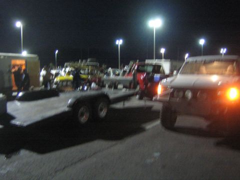 1990 Ford Bronco - empty tandem-axle flatbed trailer beside a light-colored SUV with headlights on in a parking lot at night.