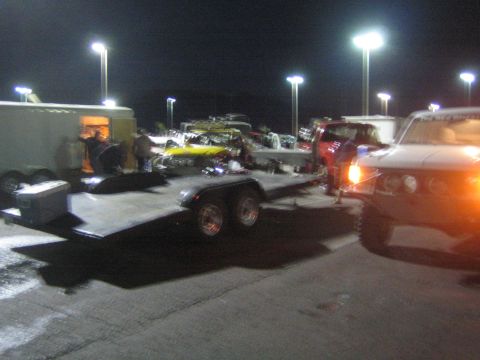 1990 Ford Bronco - flatbed trailer staged near off-road race vehicles and an enclosed trailer with a tow SUV at night.
