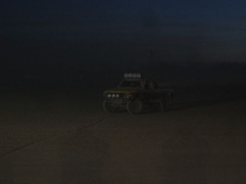 1990 Ford Bronco - off-road race truck with a roof light bar drives across open desert terrain in low dusk light.