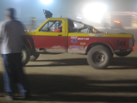 1990 Ford Bronco - red and yellow number 744 off-road race truck kicks up dust on a dirt course at night as a man watches.
