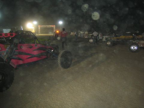 1990 Ford Bronco - pink and black tube-frame off-road buggy among other race vehicles on a dusty dirt course at night.