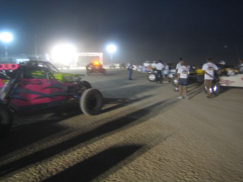 1990 Ford Bronco - pink tube-frame off-road buggy and crew members on a floodlit dirt staging area at night.