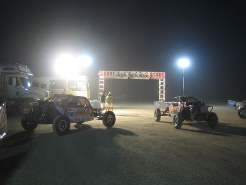 1990 Ford Bronco - Two tube-frame off-road race buggies staged on dirt near a lit SCORE start arch at night.