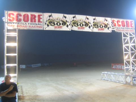 1990 Ford Bronco - SCORE International Off-Road Racing start arch with Laughlin 300 banners lit up over a dirt course at night.