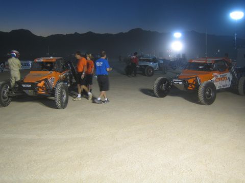 1990 Ford Bronco - Two orange tube-frame off-road race buggies and crew on a dirt staging area against mountains at dusk.