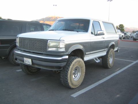 1990 Ford Bronco - two-tone white-and-gray Ford Bronco with a lift kit and large off-road tires sits in a paved parking lot at dusk.