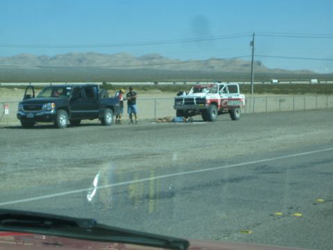 1990 Ford Bronco - dark pickup truck tows a battered red-and-white off-road prerunner truck on a paved shoulder beside a desert highway.